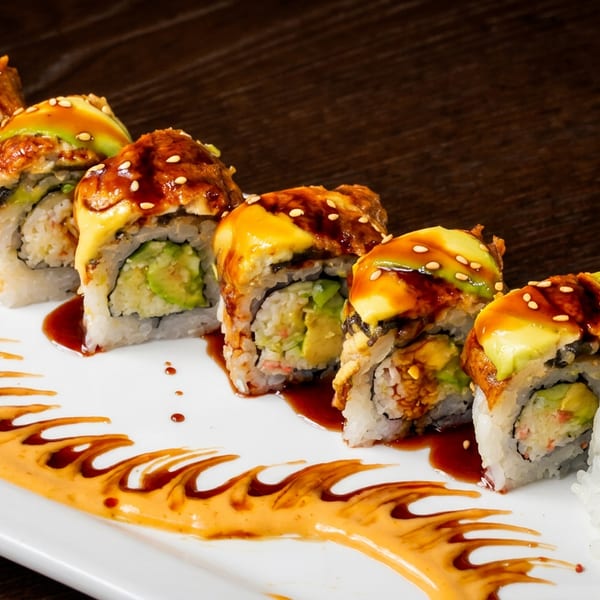Dragon Roll sushi with avocado, eel, sesame seeds, and sauces, on a white plate.